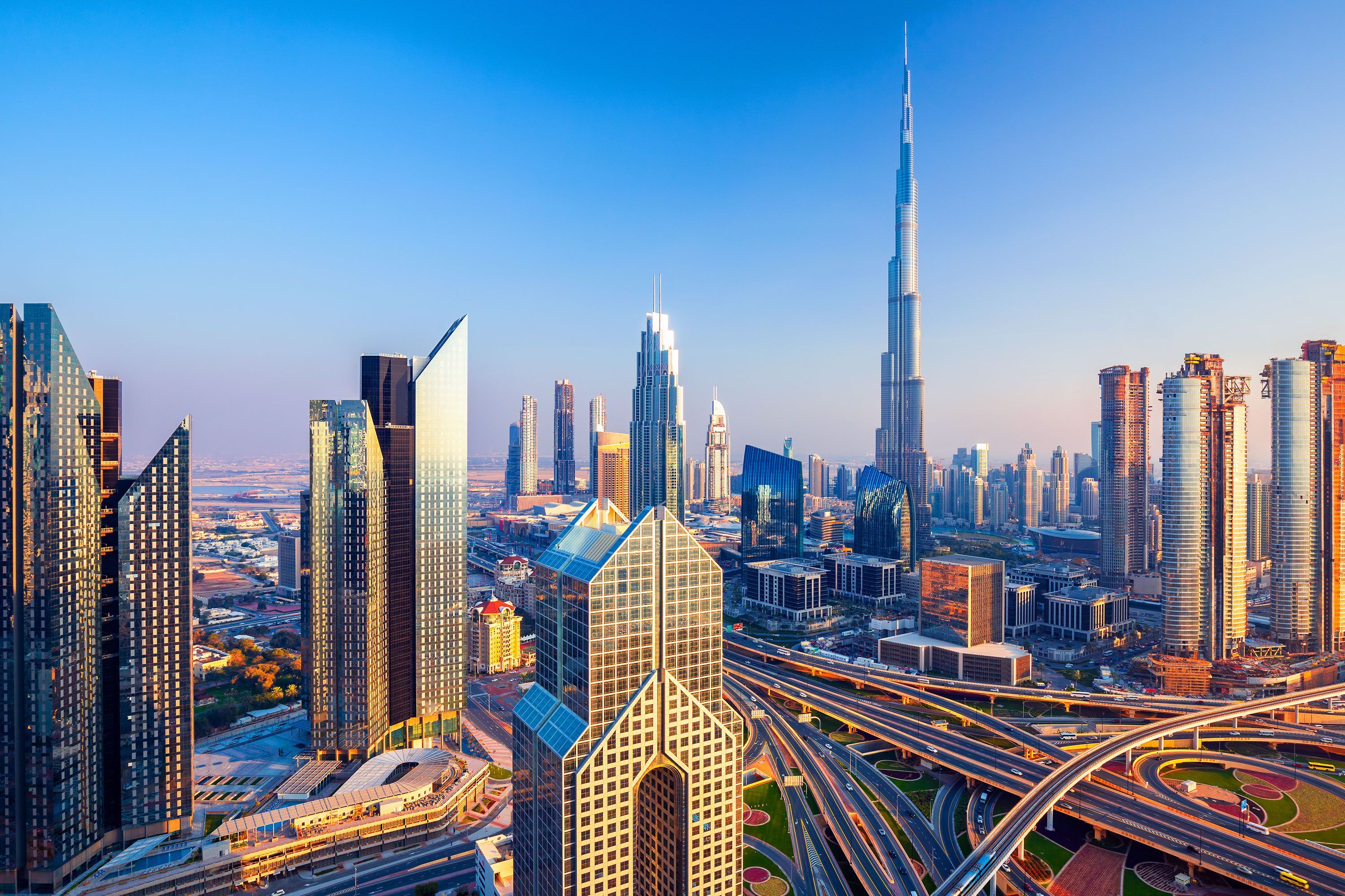 Dubai city center skyline at sunset, with a mix of modern high-rise buildings illuminated in warm orange and pink hues against a darkening sky.