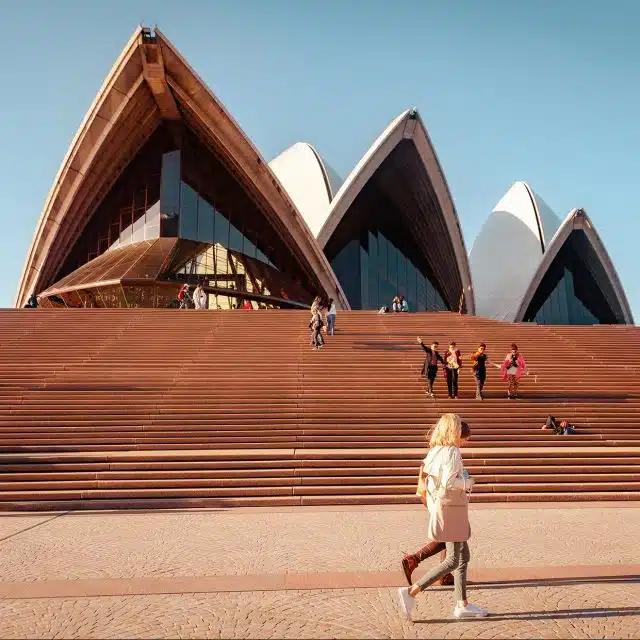 People walk by and others sit on the stairs at the front of the landmark Sydney Opera House in Sydney, Australia
