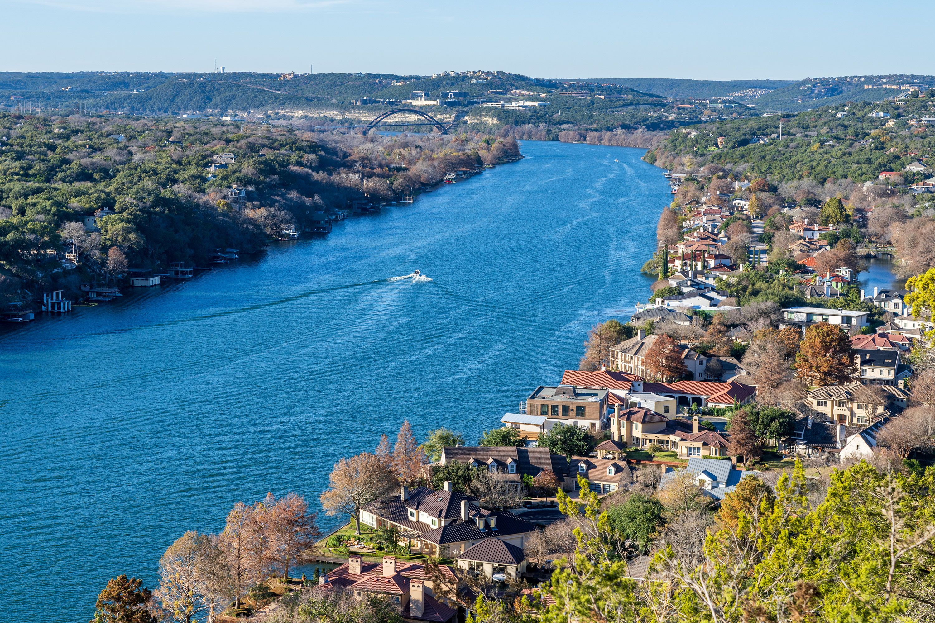 A winding river flows past hillside homes and greenery under a clear blue sky.