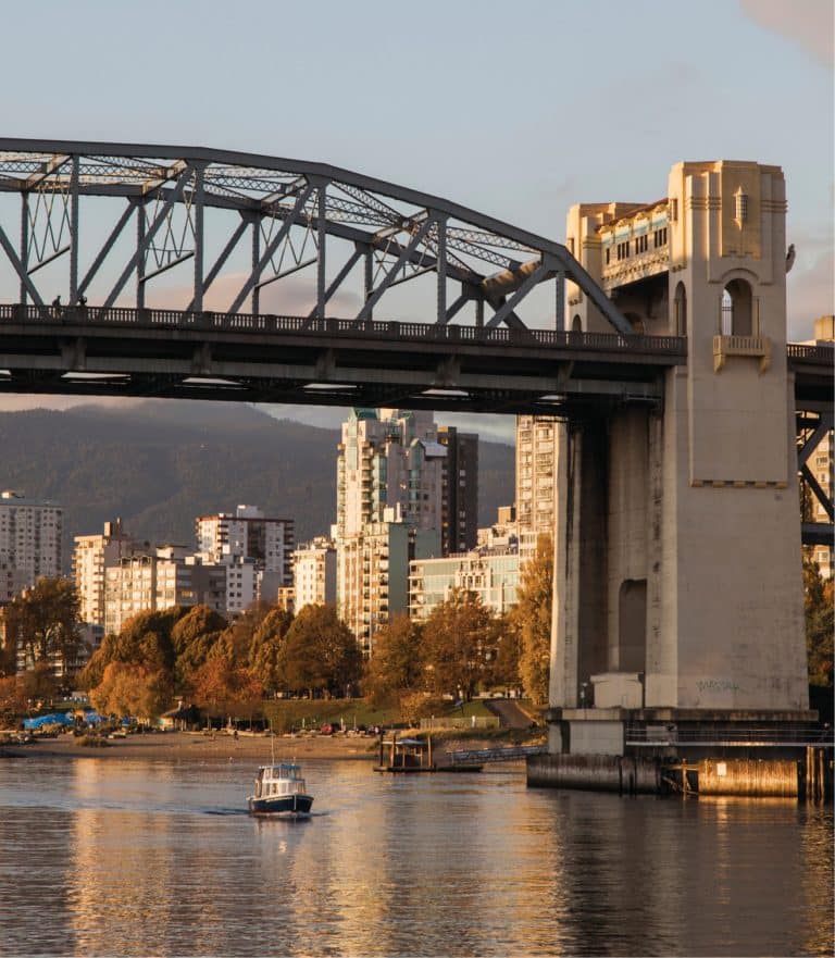 Vancouver's Burrard Street Bridge, which spans False Creek, list in evening sunlight, with a tiny boat passing underneath