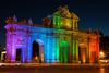 Neoclassical Alcala Gate monument in Madrid glowing with rainbow-colored lights at night.