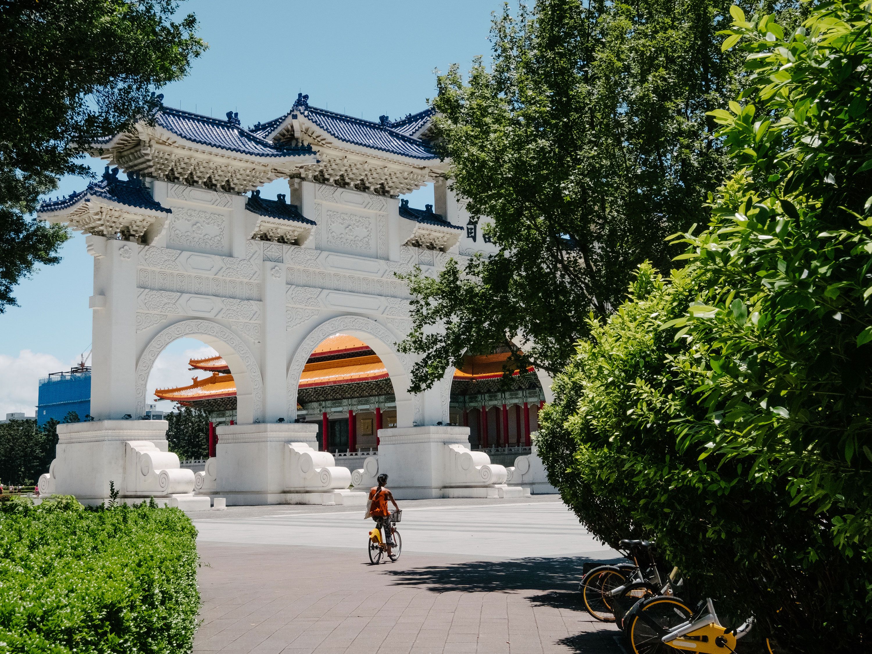 A woman riding a bike in front of a large white temple entrance surrounded by trees.