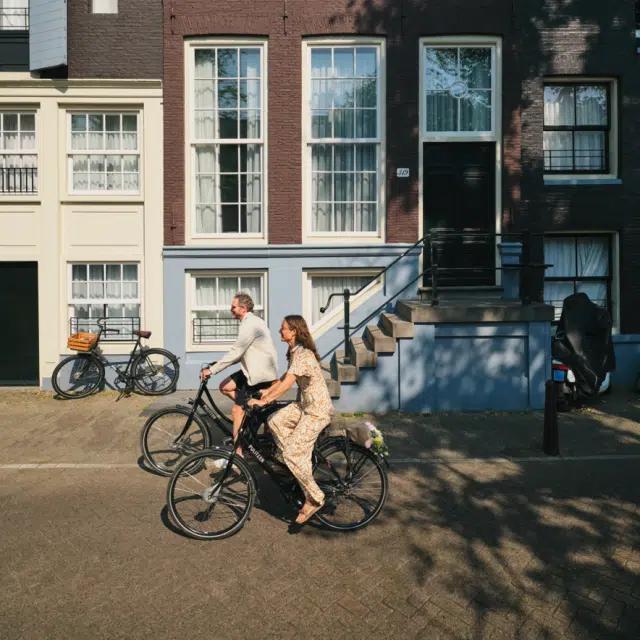 Two cyclists ride through dappled sunlight past a canal house in Amsterdam