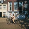 Two cyclists ride through dappled sunlight past a canal house in Amsterdam