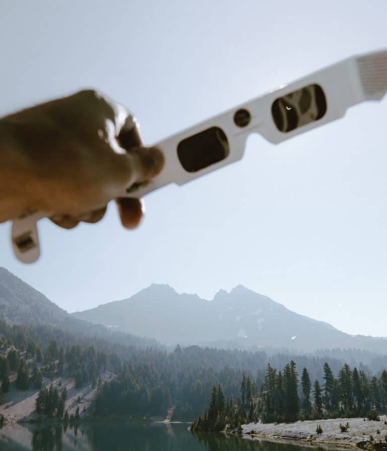 A pair of solar eclipse glasses, the lenses dark, held up to a sky, with mountains, lake and forest beyond