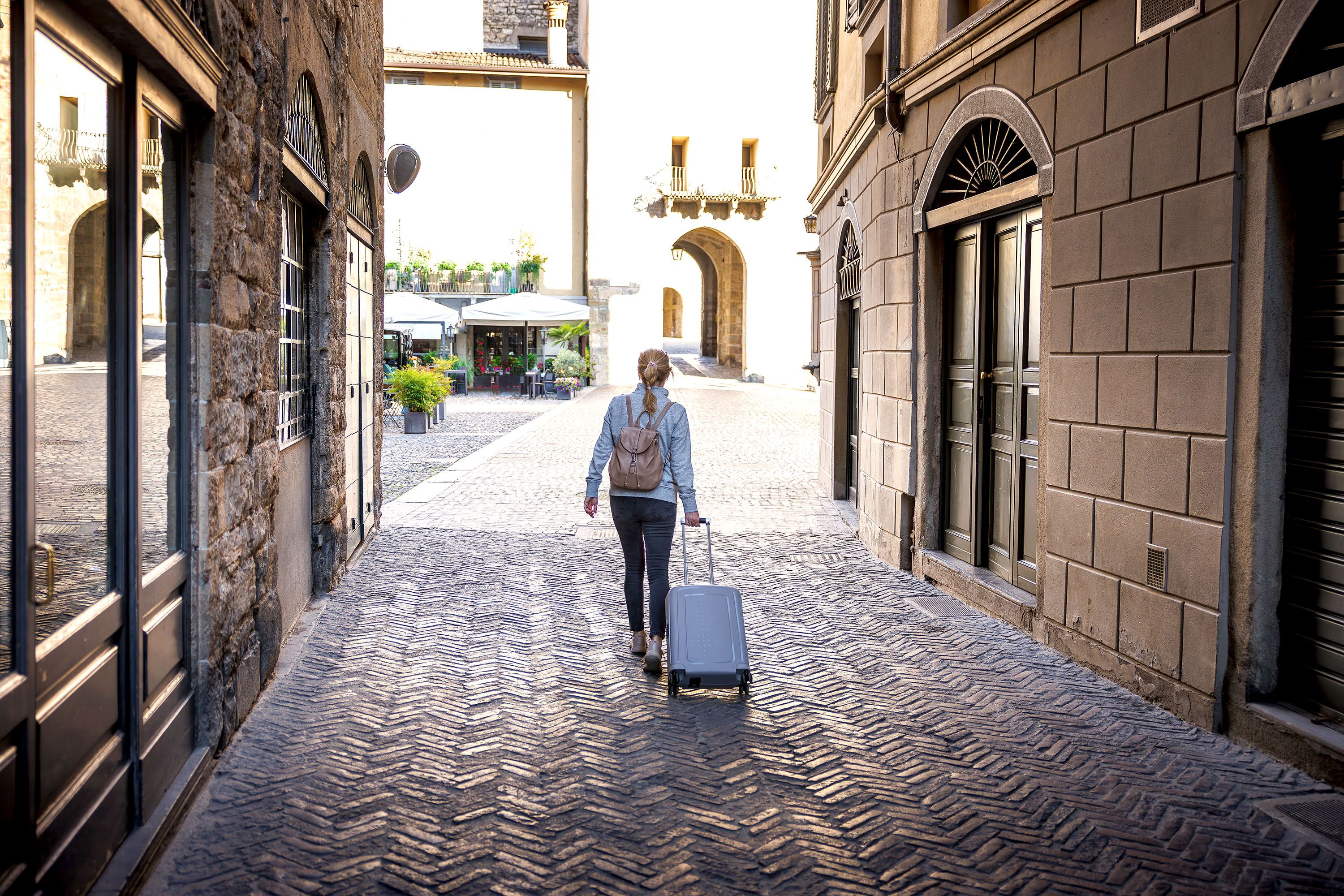 A woman walks with a suitcase through a European city with cobblestone streets.