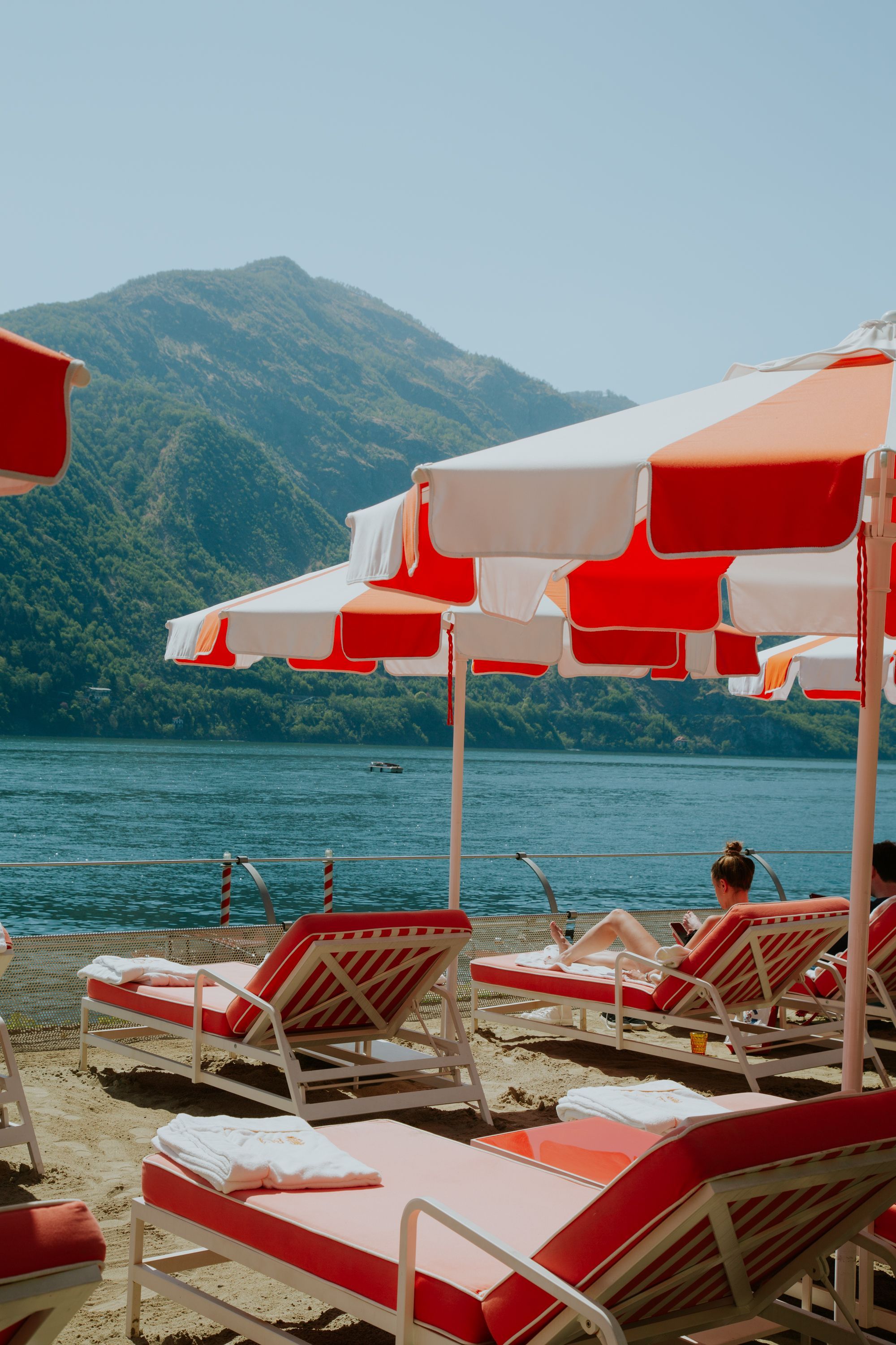 A woman sits on a red and pink sun lounger beside striped umbrellas on Lake Como, Italy