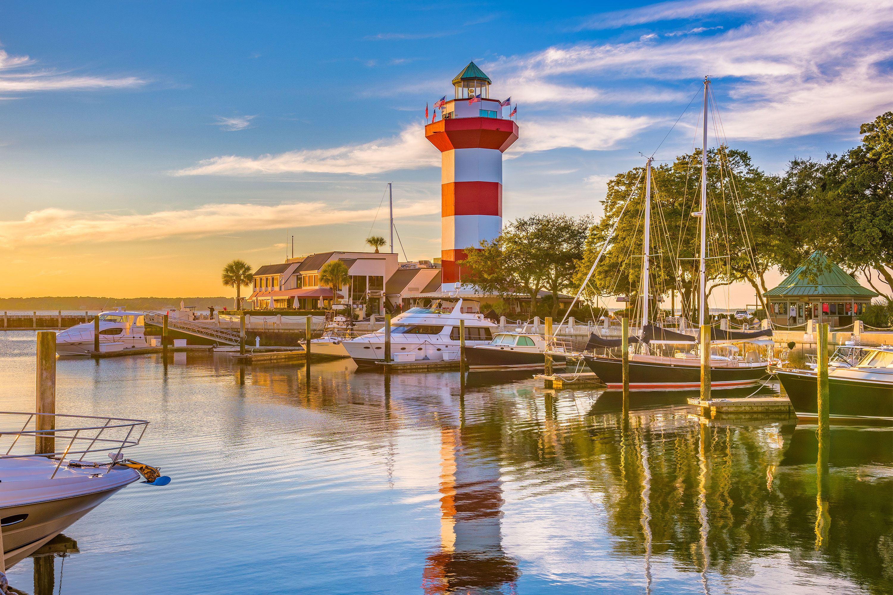 A marina scene at sunset with the red-and-white Harbour Town Lighthouse, docked boats, waterfront buildings, and trees reflected in the still water.
