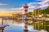 A marina scene at sunset with the red-and-white Harbour Town Lighthouse, docked boats, waterfront buildings, and trees reflected in the still water.