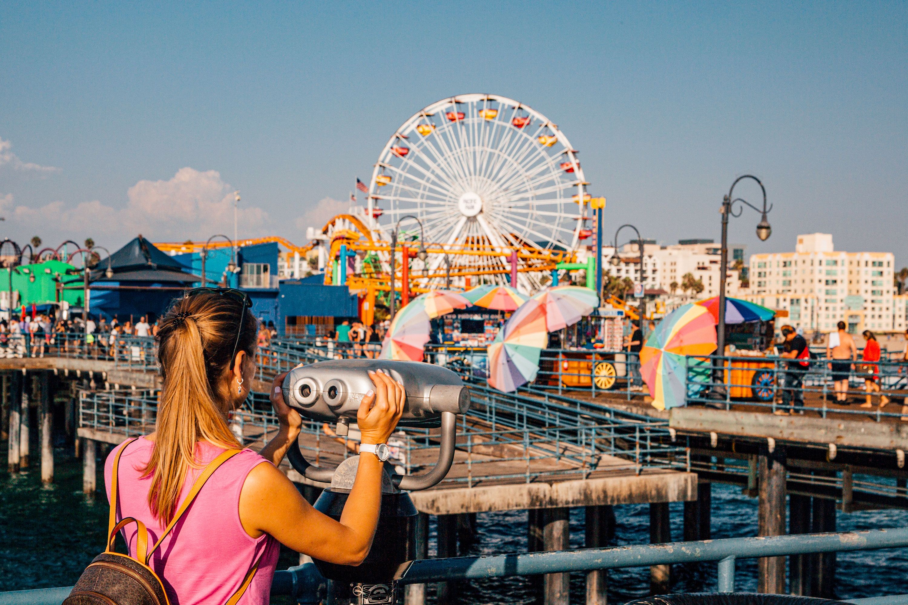 A woman in a pink sleeveless shirt with her hands on a tower telescope viewer looks out toward the Santa Monica pier with its Ferris wheel in Los Angeles in afternoon golden light.