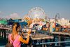 A woman in a pink sleeveless shirt with her hands on a tower telescope viewer looks out toward the Santa Monica pier with its Ferris wheel in Los Angeles in afternoon golden light.