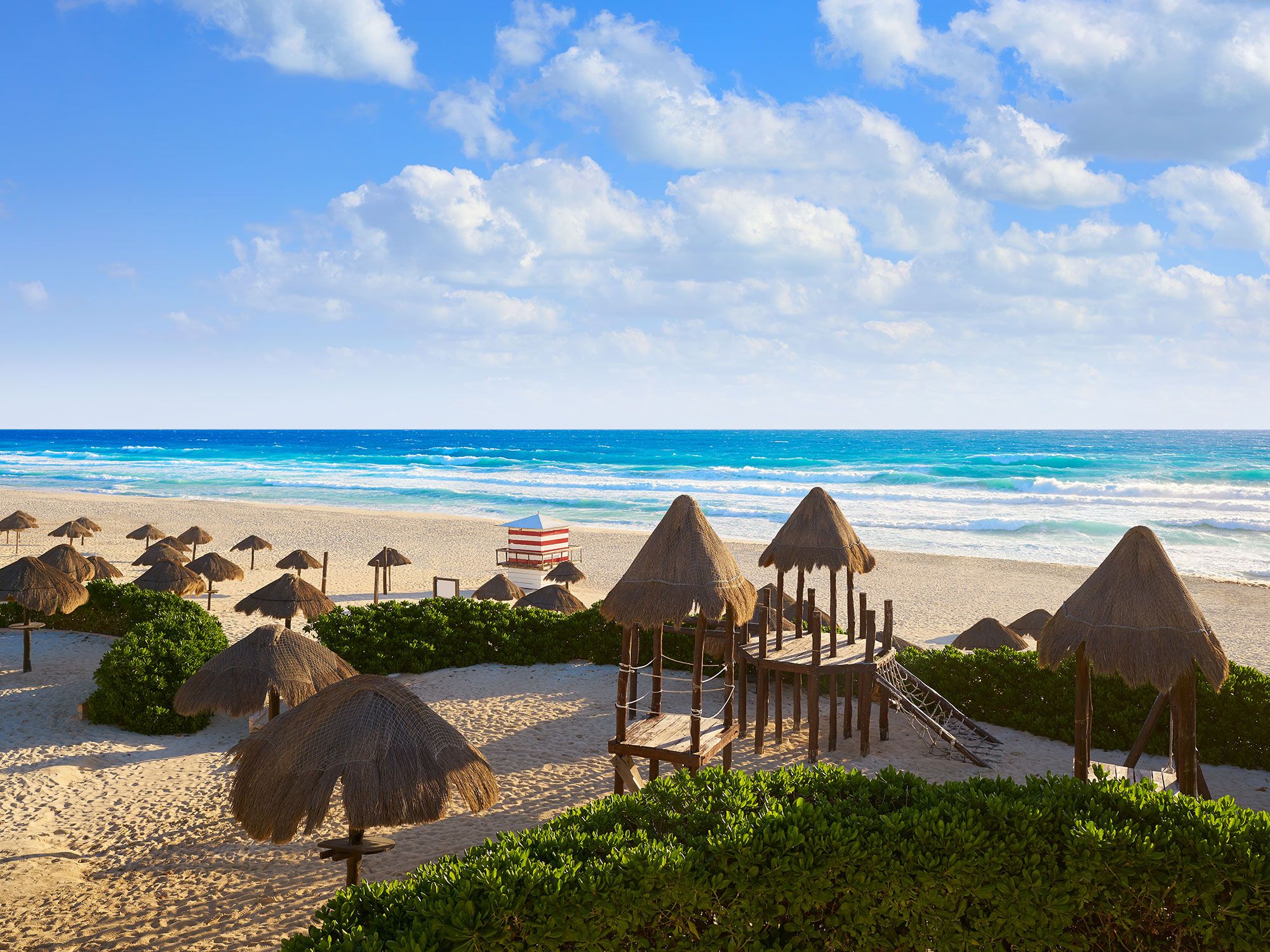 Sea grass umbrellas line a beach with a small red and white lined shelter with the ocean in the distance.