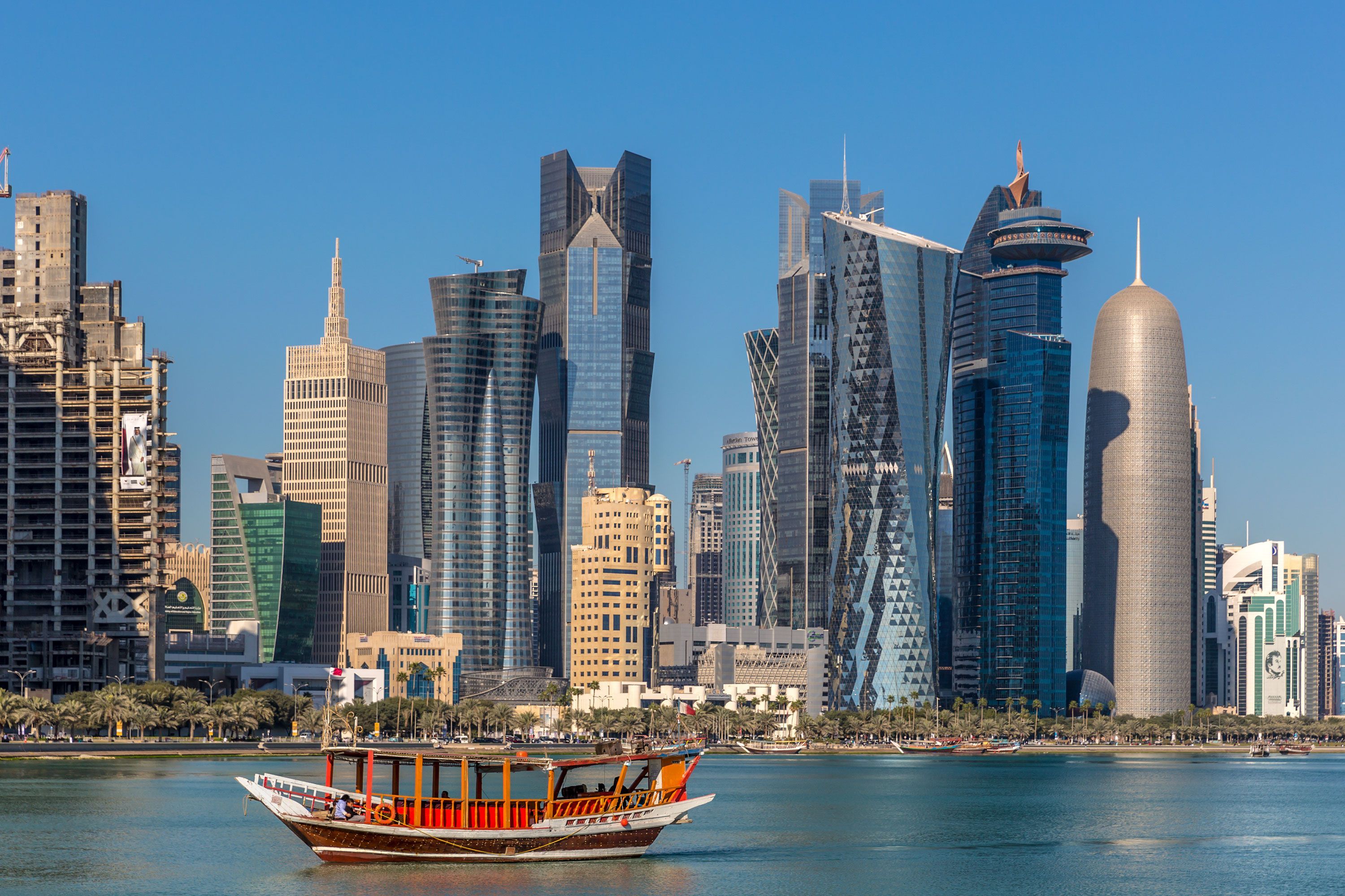 A small wooden boat on the water of Doha Bay with the glass and steel high-rise skyline of West Bay in the background under a clear blue sky.