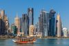 A small wooden boat on the water of Doha Bay with the glass and steel high-rise skyline of West Bay in the background under a clear blue sky.
