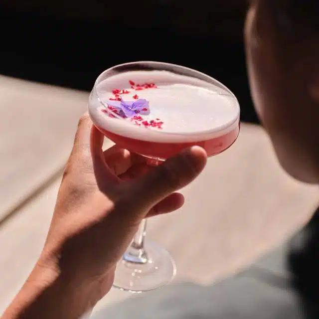 A hand raises a coupe glass containing a pink cocktail scattered with small edible flower petals