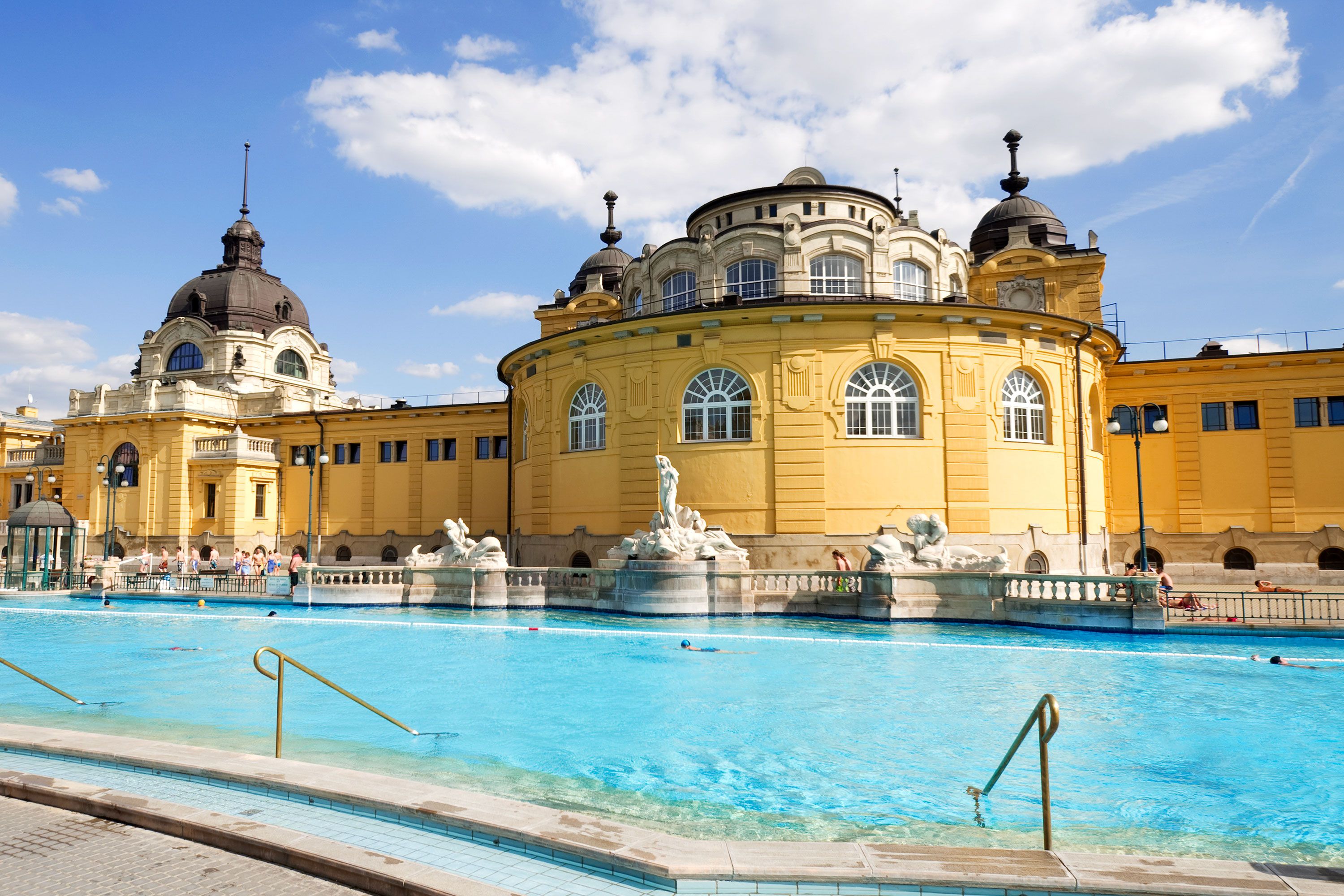 A wide exterior view of the Széchenyi Thermal Bath in Budapest, showing its pale-yellow Neo-Baroque building, domed towers, and outdoor pools surrounded by terraces on a sunny day.