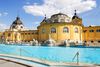 A wide exterior view of the Széchenyi Thermal Bath in Budapest, showing its pale-yellow Neo-Baroque building, domed towers, and outdoor pools surrounded by terraces on a sunny day.