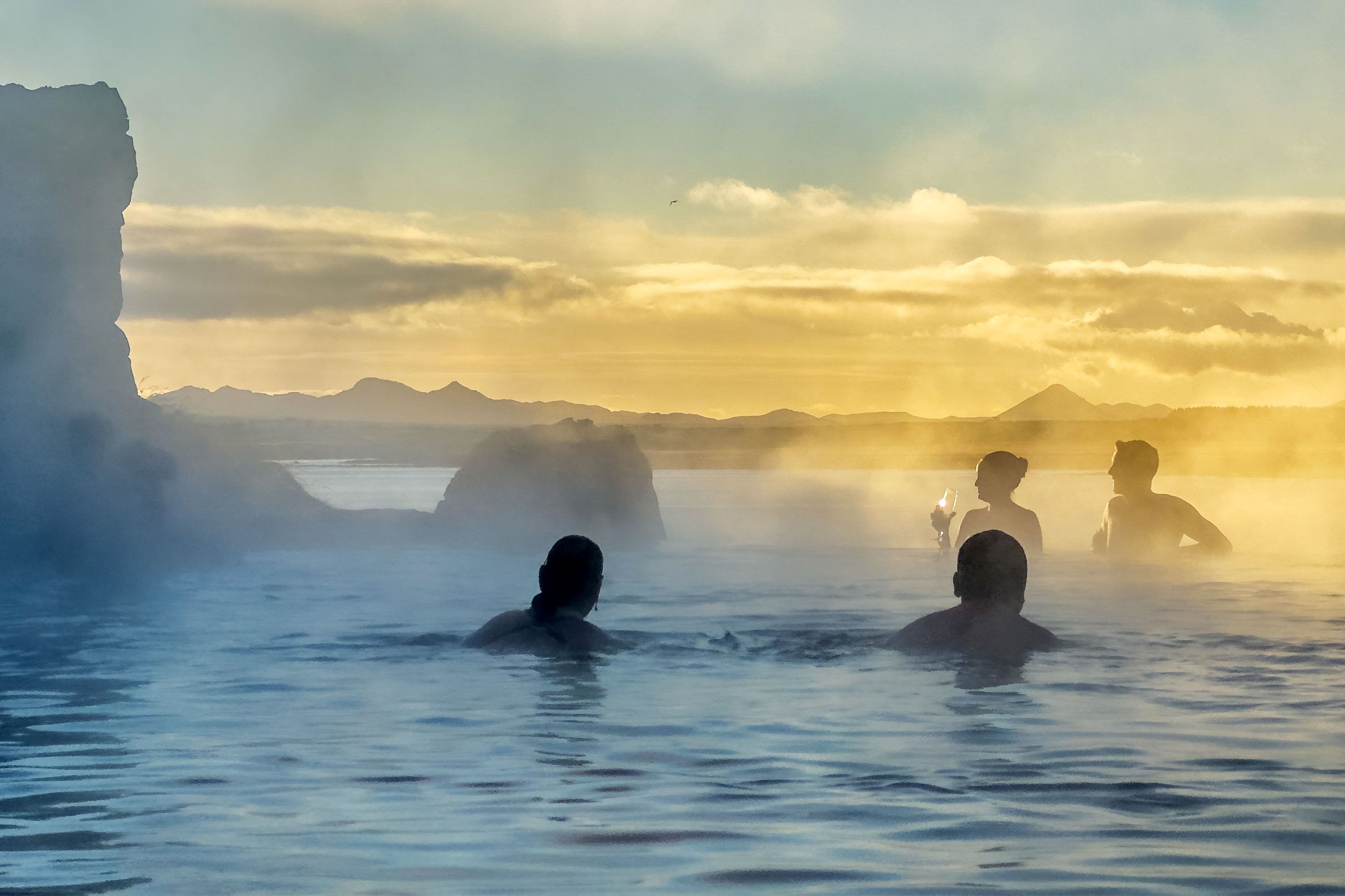 People relaxing in the geothermal infinity-edge pool at Sky Lagoon in Iceland, with steam rising over warm water and a clear sky above the ocean.