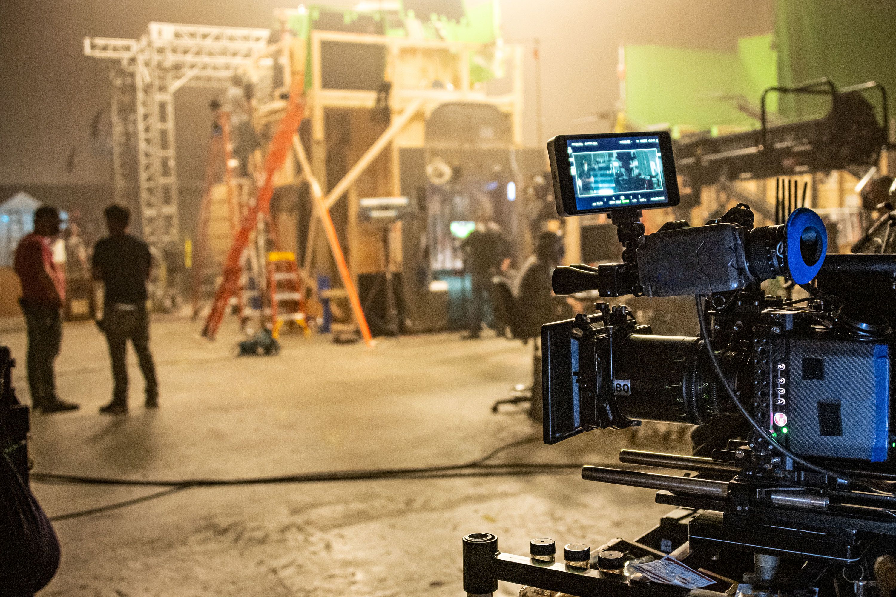 With a professional movie camera in the right foreground of the frame an out-of-focus scene on the set of a movie production.