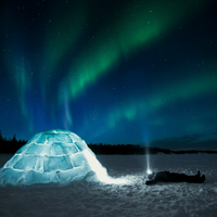 A person lies in the snow next to an illuminated igloo looking up at the northern lights lighting up the sky above