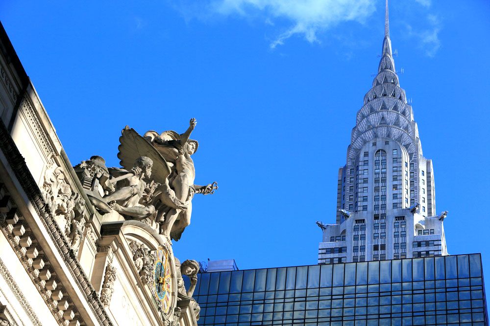 Statues and carvings on top of the Grand Central Sation entrance, the Empire State Building tower in the background.