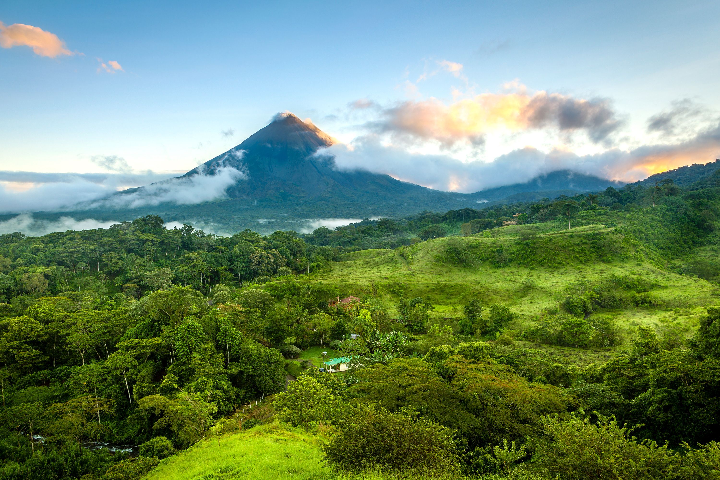 A lush, green valley leads to a symmetrical volcanic peak partially shrouded by drifting clouds.