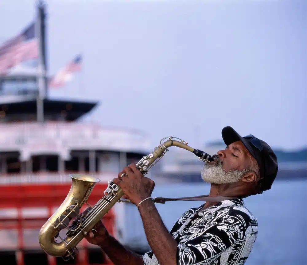 A New Orleans musician wearing a baseball cap and patterned black and white shirt plays a saxophone, with a view of a boat in the Mississippi River behind him