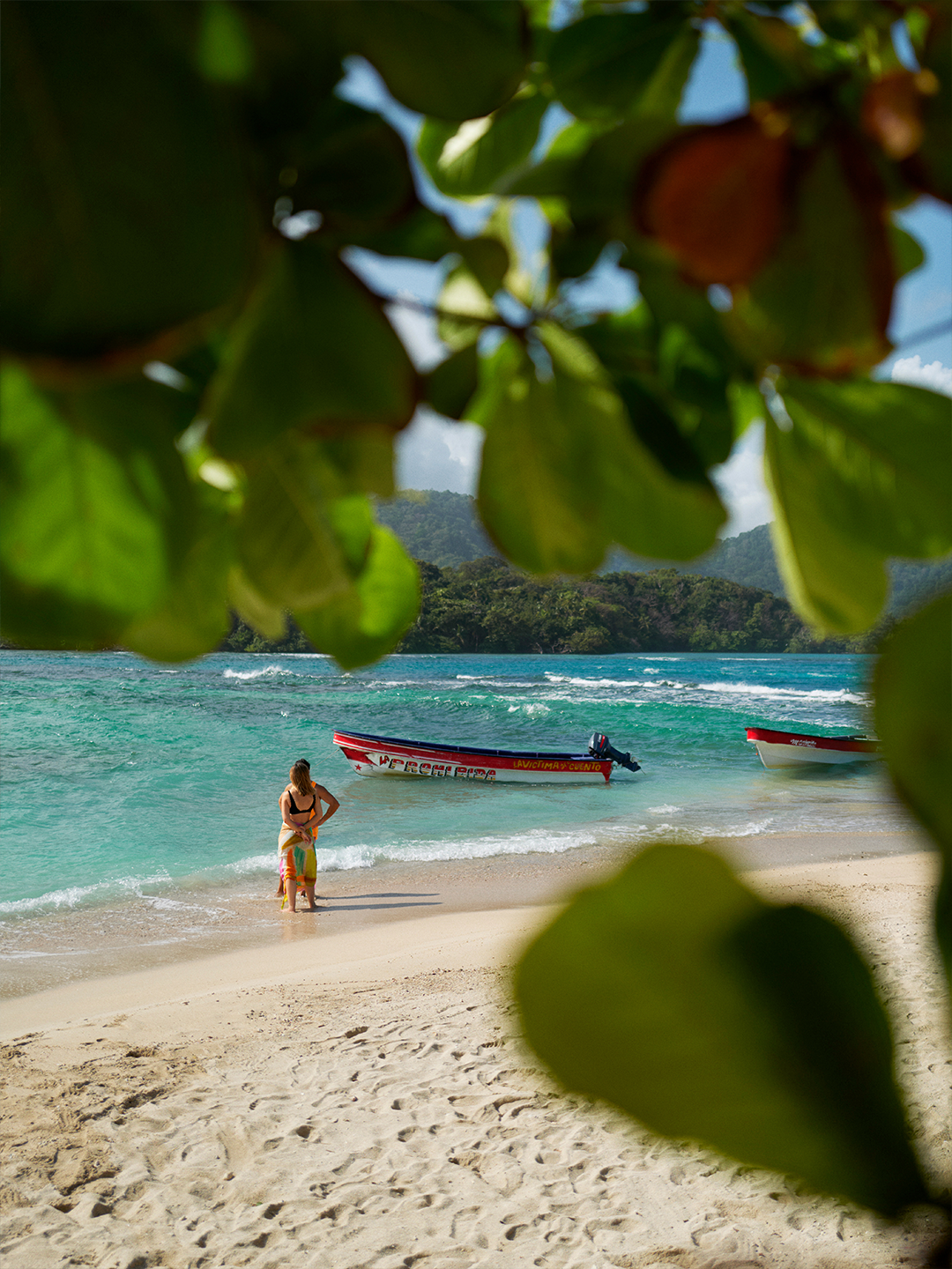 A person stands on a sandy beach in Panama looking out at the blue ocean, seen between green leaves 