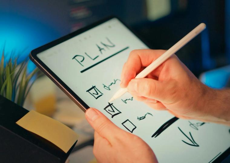 Hands using a stylus to check off items on a digital 'PLAN' checklist on a tablet.