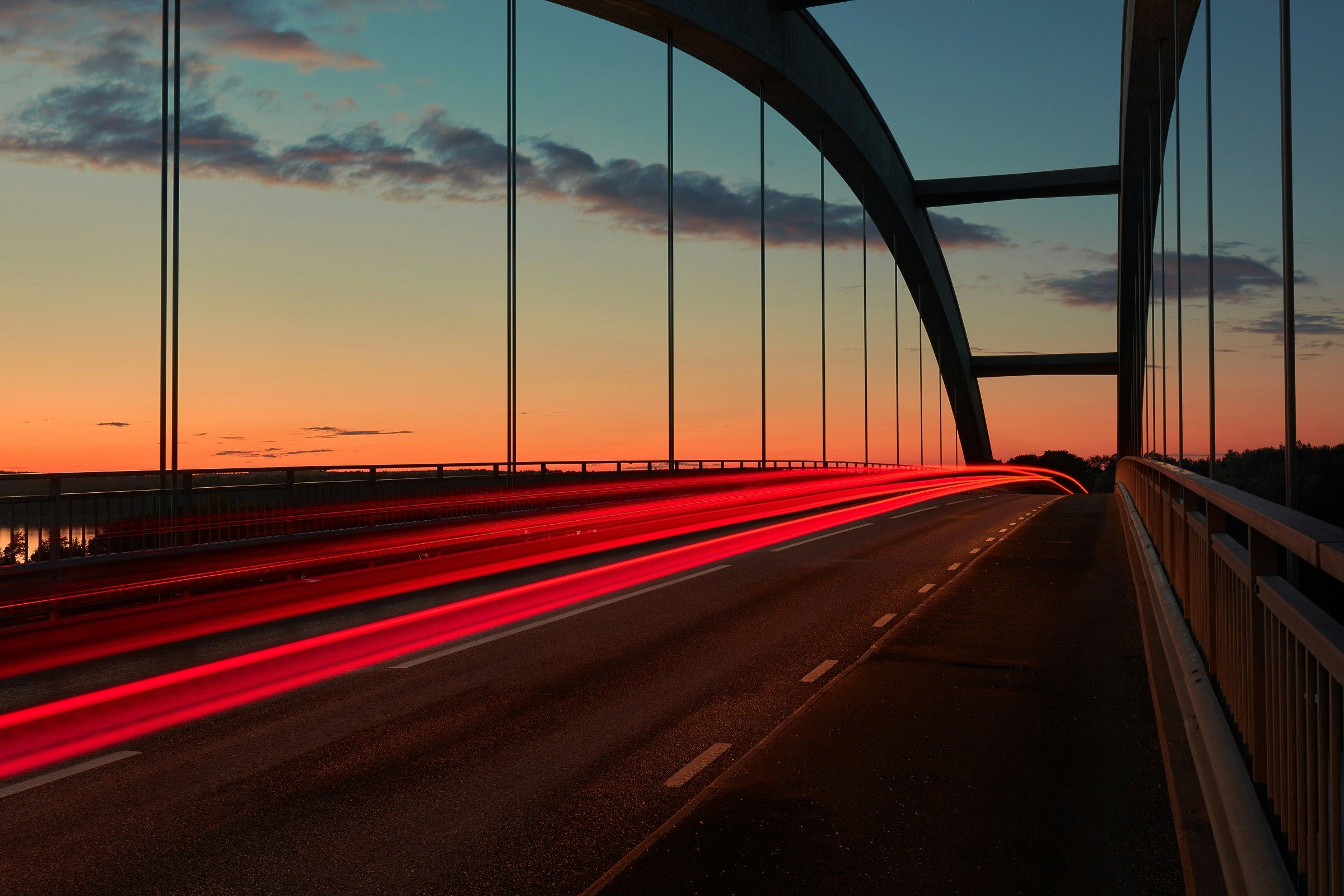 Red light trails streak across an arch bridge at sunset.