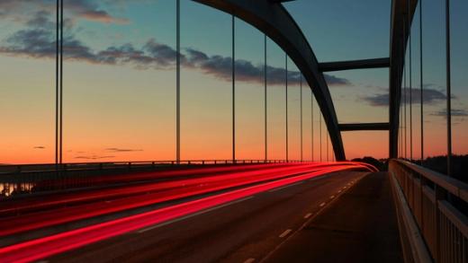 Red light trails streak across an arch bridge at sunset.