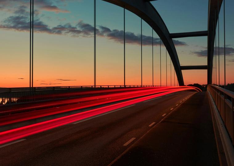 Red light trails streak across an arch bridge at sunset.