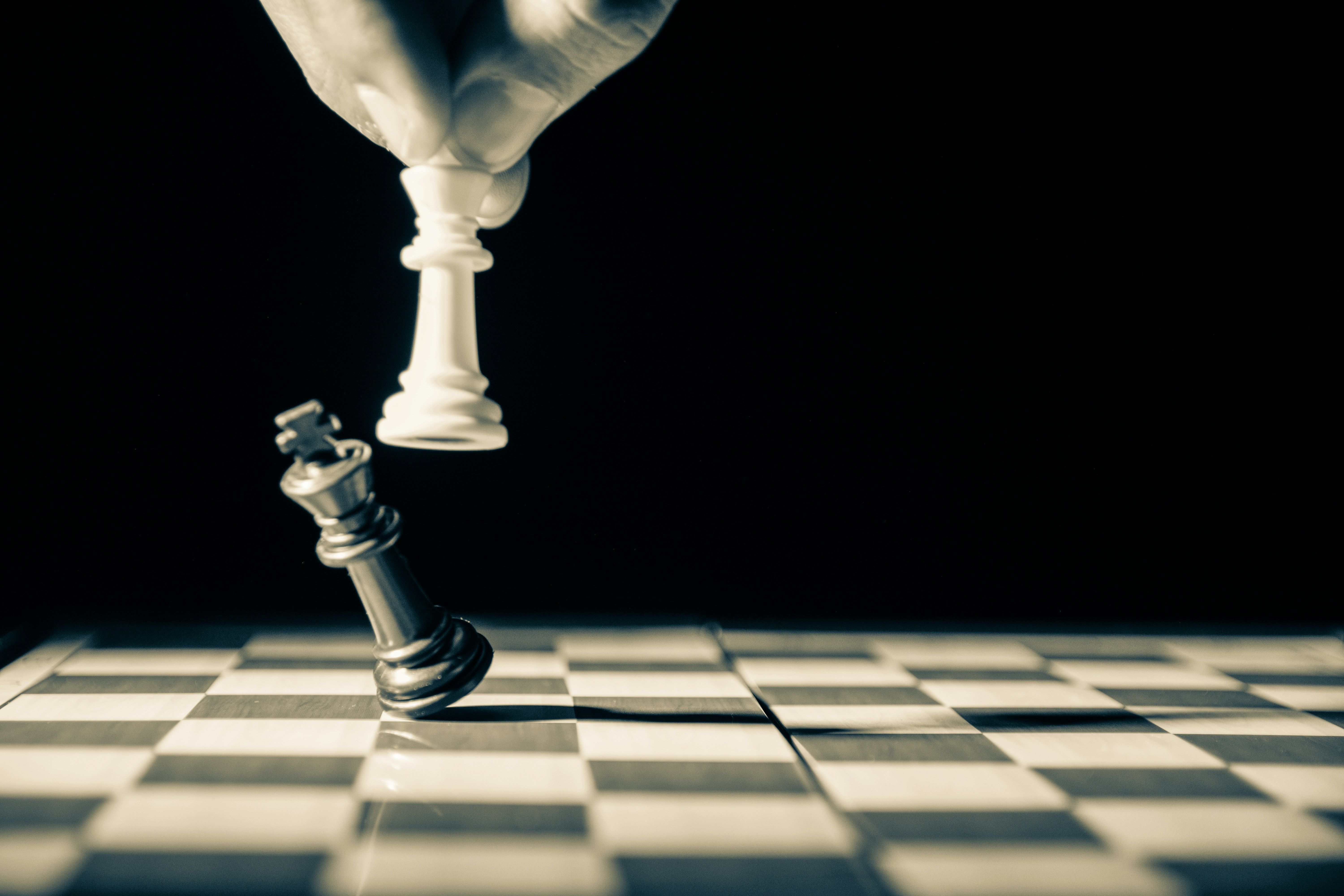 A hand holds a white chess king above a falling silver king on a chessboard.