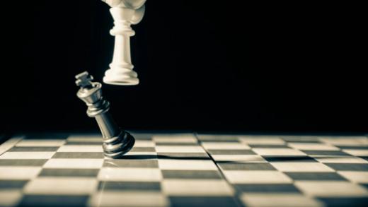 A hand holds a white chess king above a falling silver king on a chessboard.