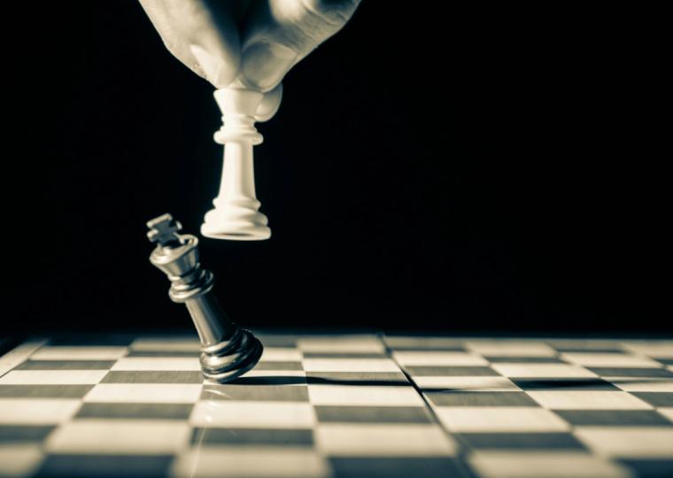 A hand holds a white chess king above a falling silver king on a chessboard.