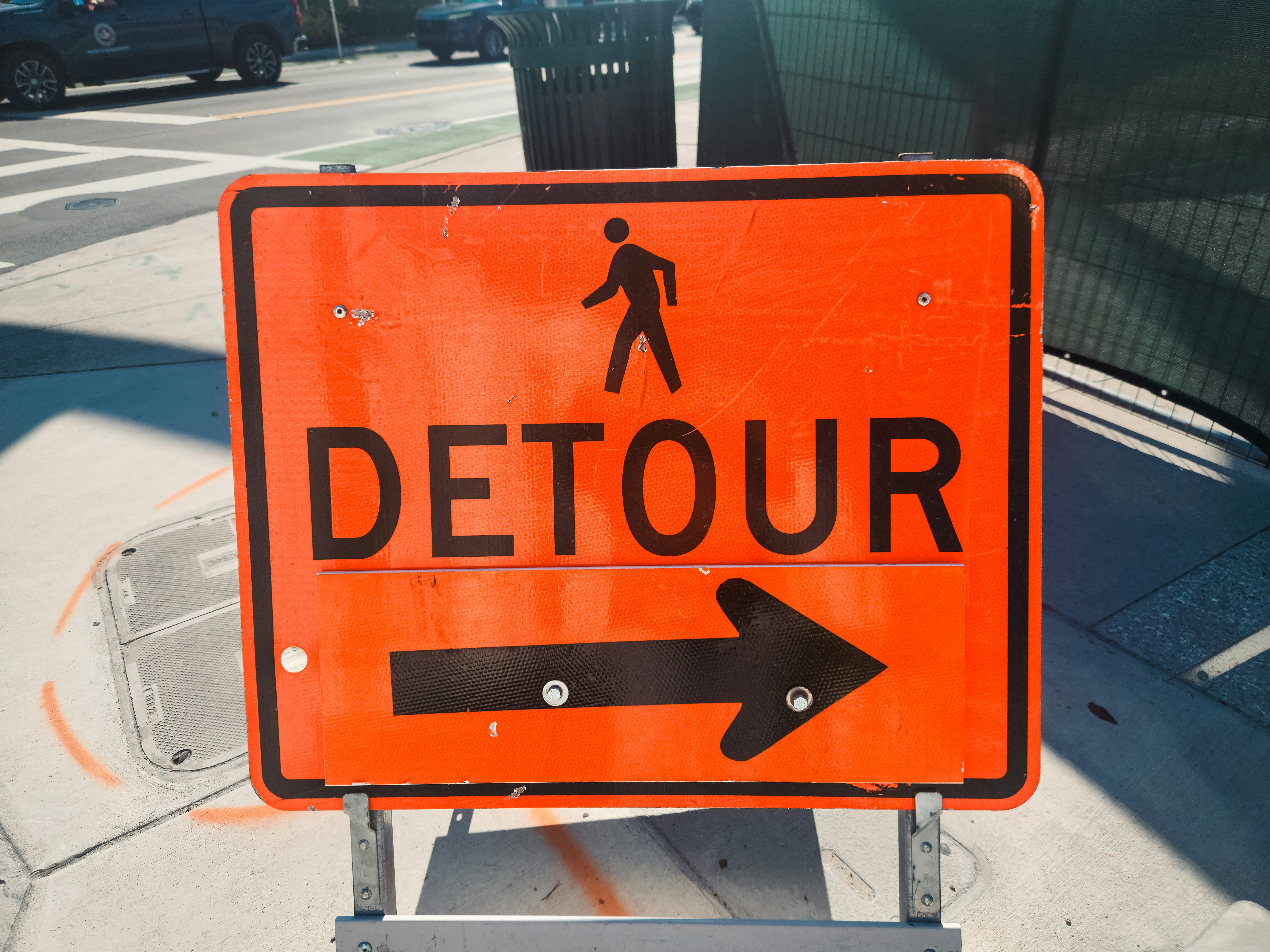 An orange sign for a pedestrian detour, featuring a walking person icon, the word "DETOUR", and an arrow pointing right.