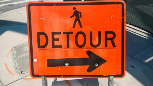 An orange sign for a pedestrian detour, featuring a walking person icon, the word "DETOUR", and an arrow pointing right.