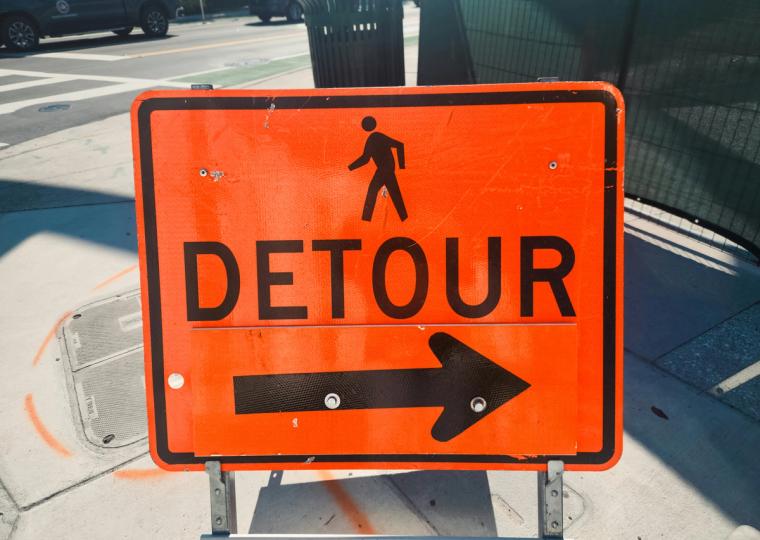 An orange sign for a pedestrian detour, featuring a walking person icon, the word "DETOUR", and an arrow pointing right.