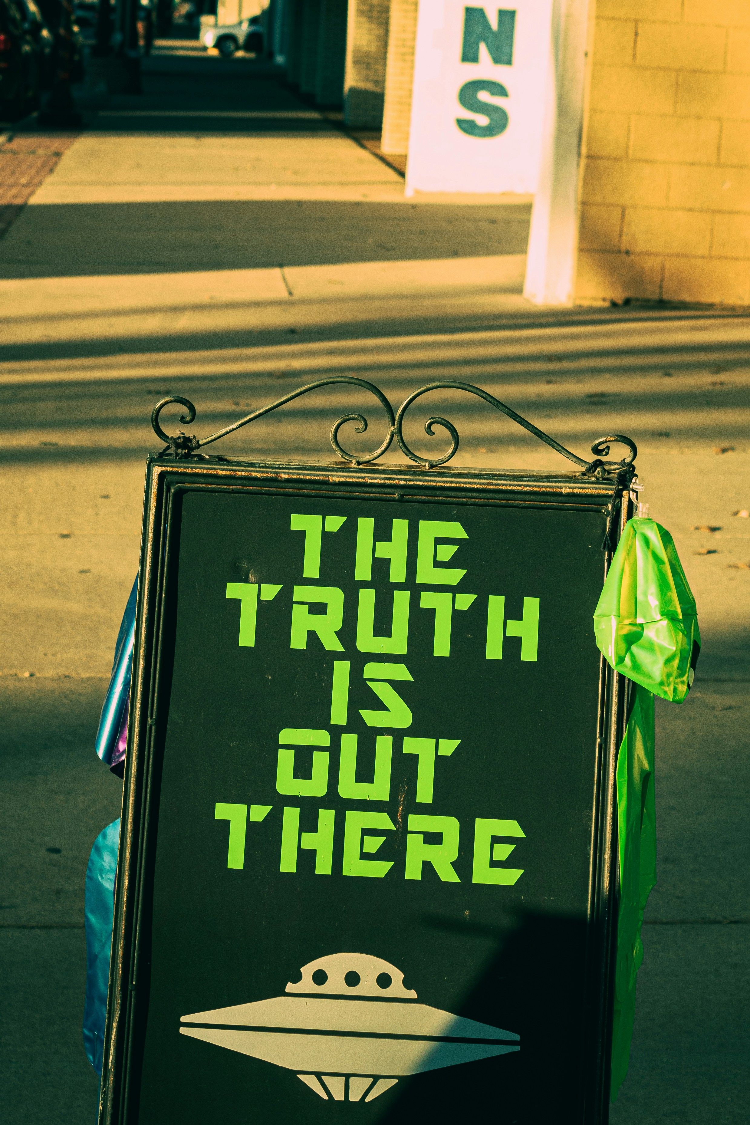 A black sidewalk sign reads "THE TRUTH IS OUT THERE" in green block letters, with a white flying saucer graphic below, and green alien bags hanging from its side.