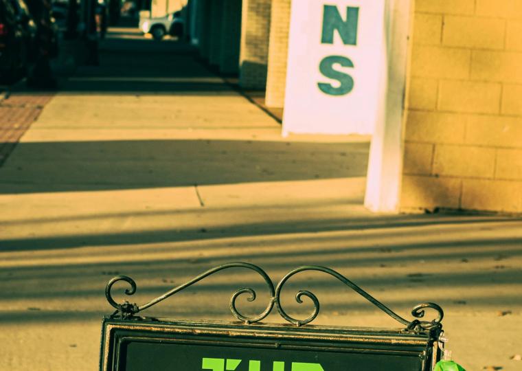 A black sidewalk sign reads "THE TRUTH IS OUT THERE" in green block letters, with a white flying saucer graphic below, and green alien bags hanging from its side.