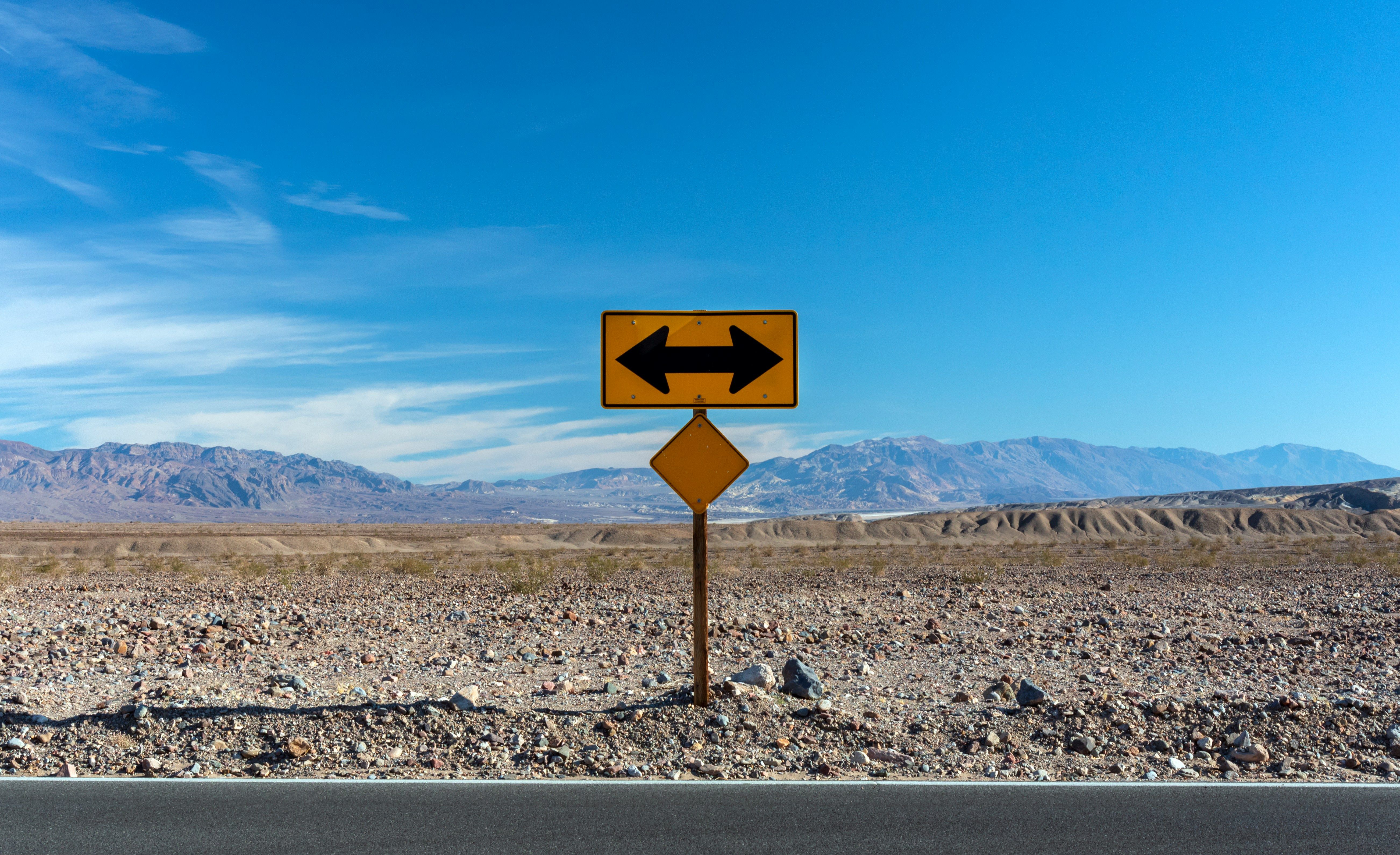 A yellow sign with opposing arrows stands in a vast desert landscape under a blue sky.