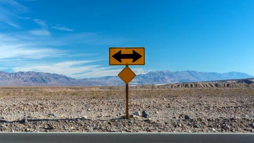 A yellow sign with opposing arrows stands in a vast desert landscape under a blue sky.