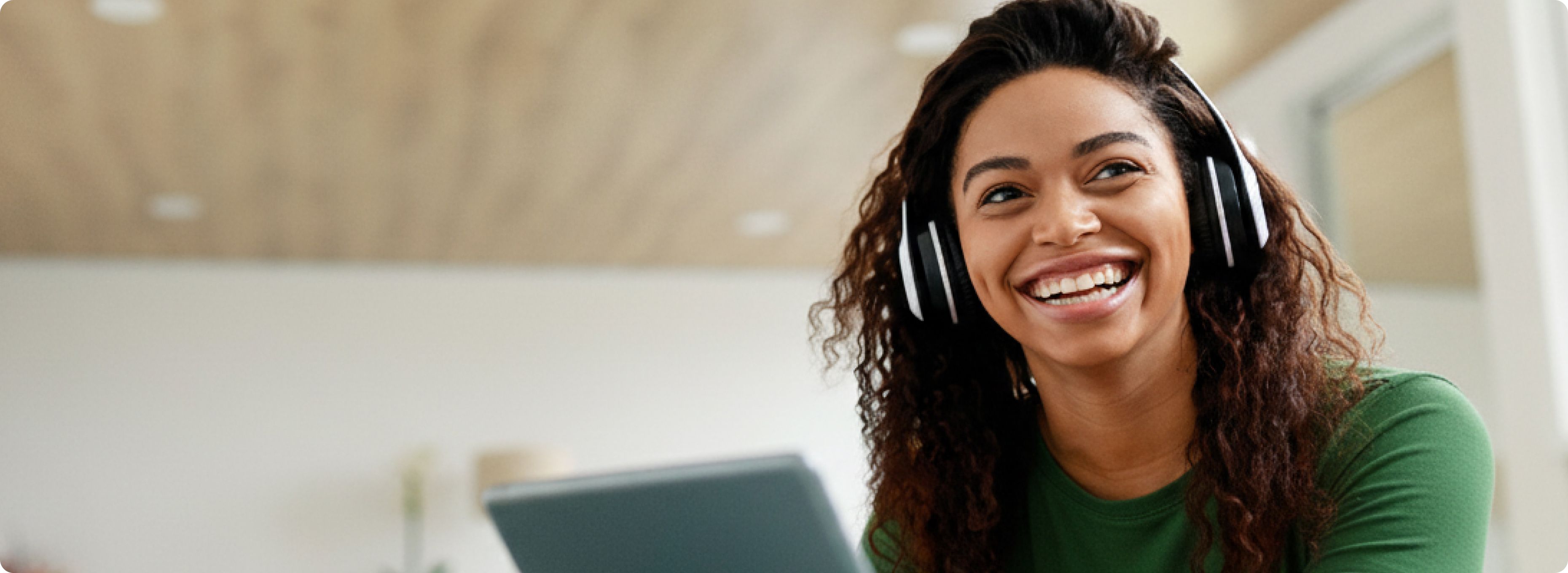 Smiling woman wearing headphones and a green shirt looks to the side while holding a laptop. She is seated in a bright room, creating a joyful atmosphere.