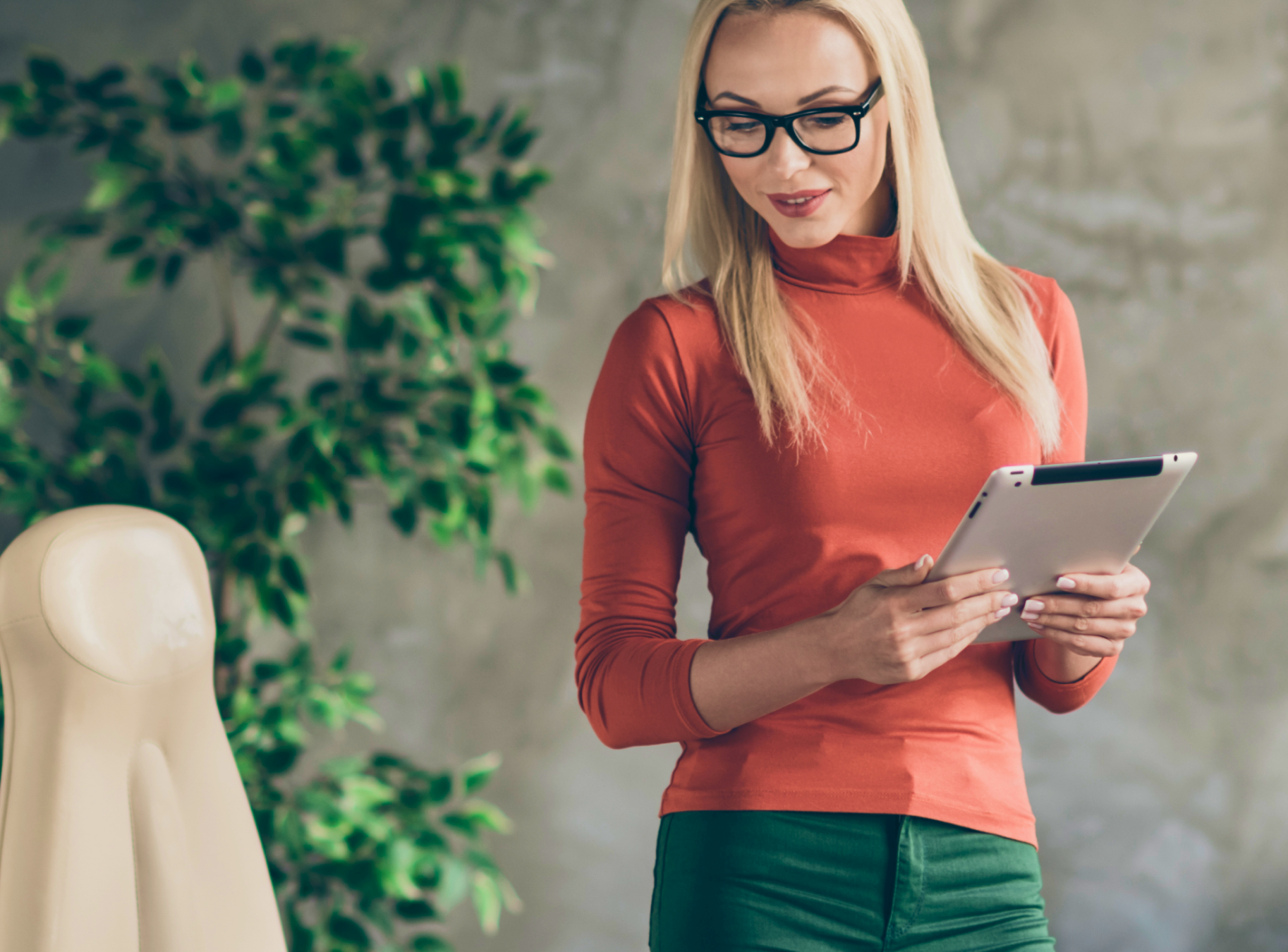 A woman with long blonde hair and glasses stands holding a tablet. She wears an orange turtleneck, exuding a relaxed, professional vibe.