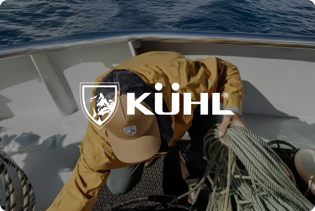 A person in a yellow jacket and cap is handling ropes on a boat. The ocean is visible in the background. The word "KÜHL" and a logo overlay the image.