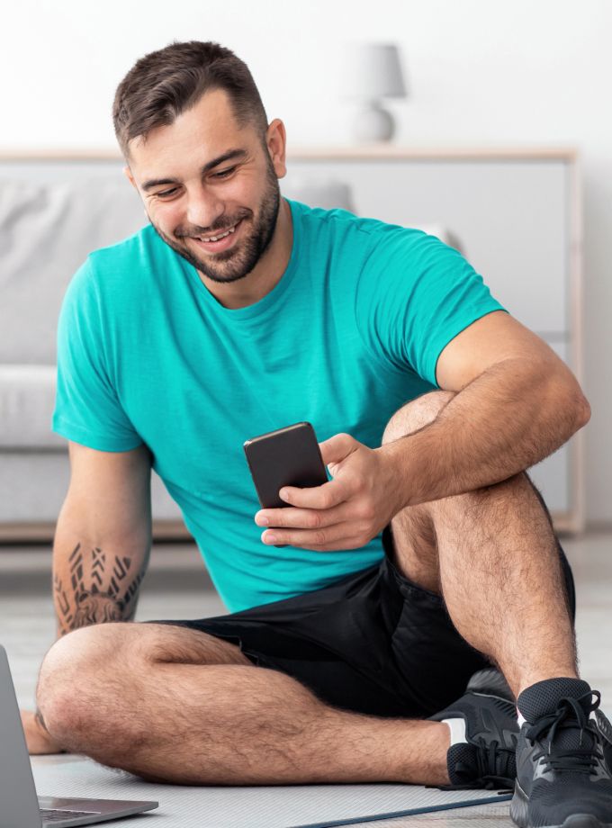A smiling man in a turquoise shirt and black shorts sits cross-legged indoors, looking at his phone. He's on a yoga mat, conveying a relaxed, casual mood.