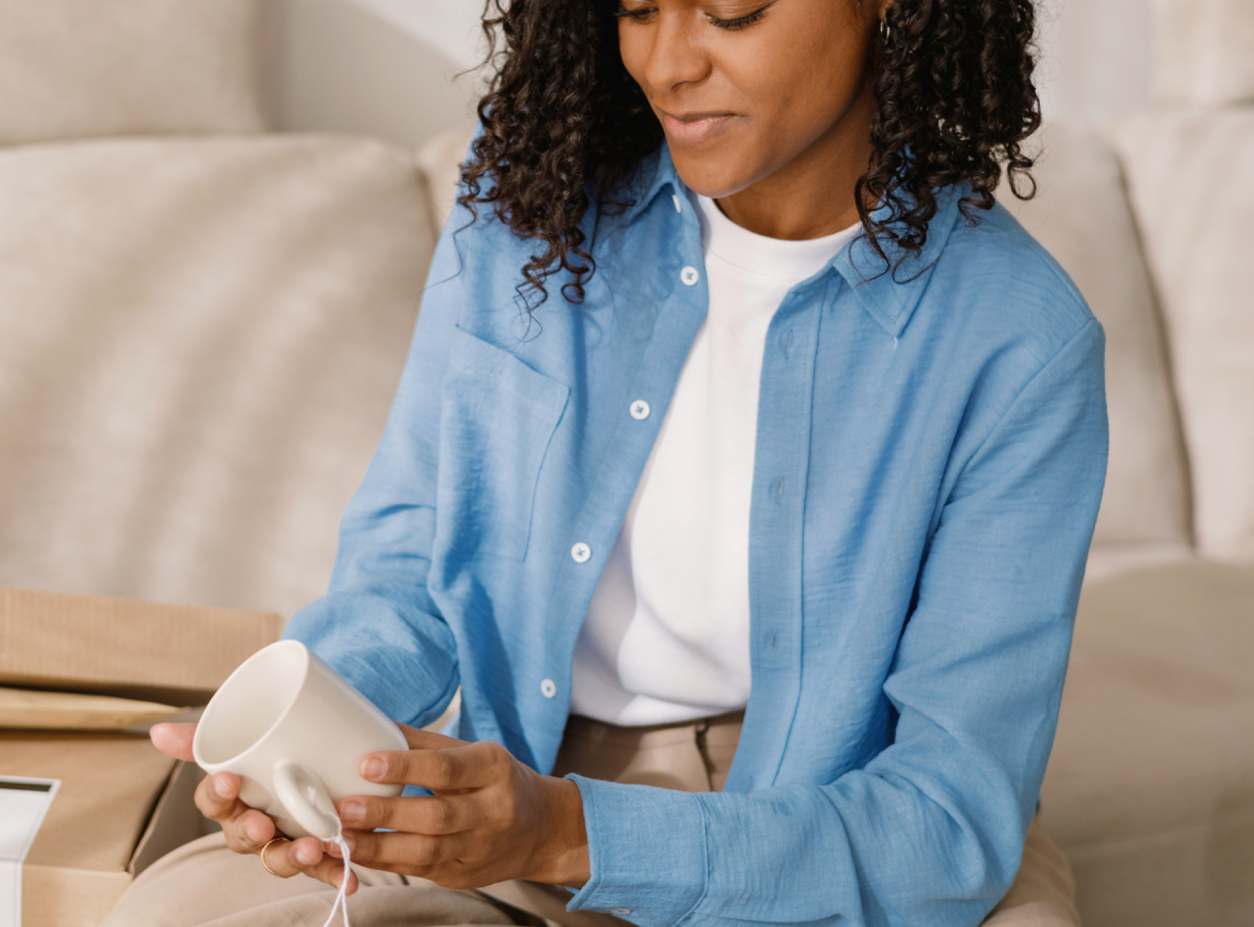 A woman in a blue shirt smiles while unboxing a white mug. She sits on a beige couch with unpacked boxes around, conveying a sense of delight and anticipation.