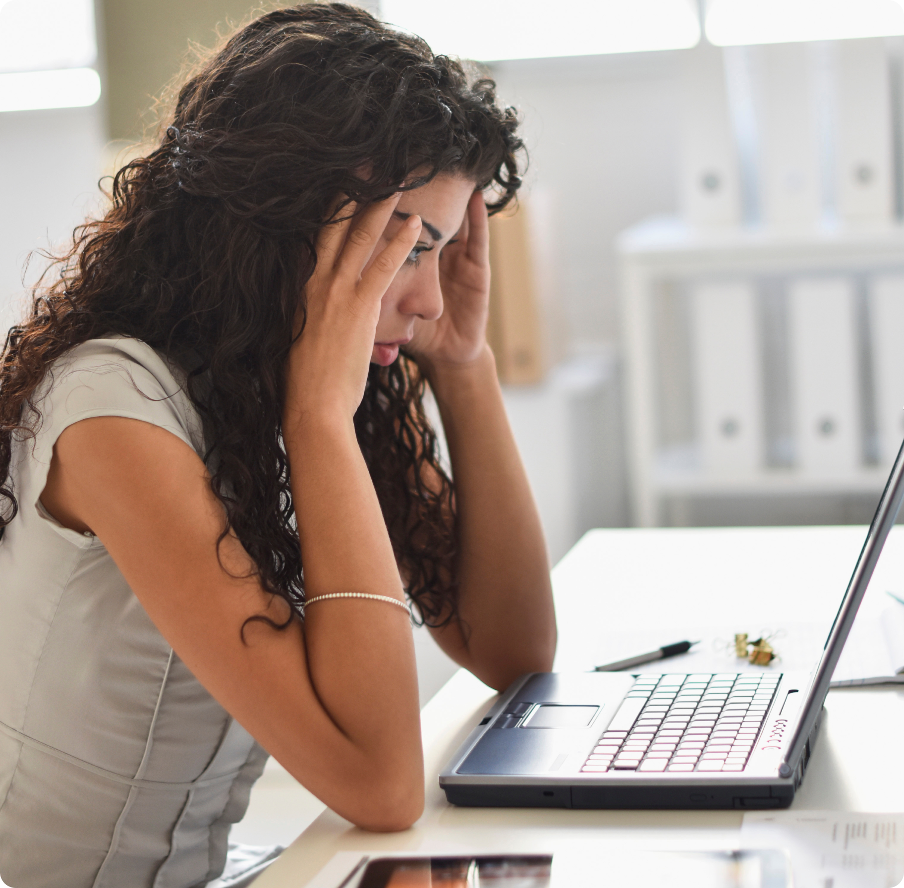 A woman with curly hair sits at a desk, looking stressed with her hands on her temples, staring at a laptop. The office setting is bright and blurred.