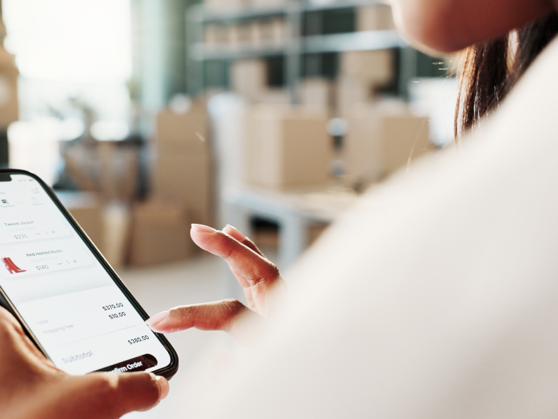 Close-up over-the-shoulder view of someone reviewing a checkout screen on their phone, with an order summary and a "confirm order" button visible, standing in a warehouse with stacked shipping boxes in the background.