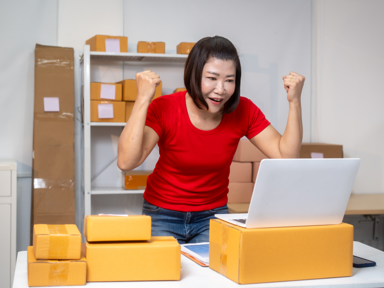 Smiling ecommerce business owner raising both fists in celebration at her laptop, surrounded by stacked shipping boxes